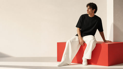 A man wearing a black short-sleeve t-shirt, white wide-leg trousers, and white sneakers sits on a bright red geometric block against a minimalist cream-colored studio background.